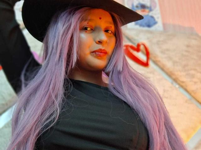 CandySwan with long purple hair, black hat, and black shirt, showing her small tits and red dot on her forehead, has a cute and confident face.