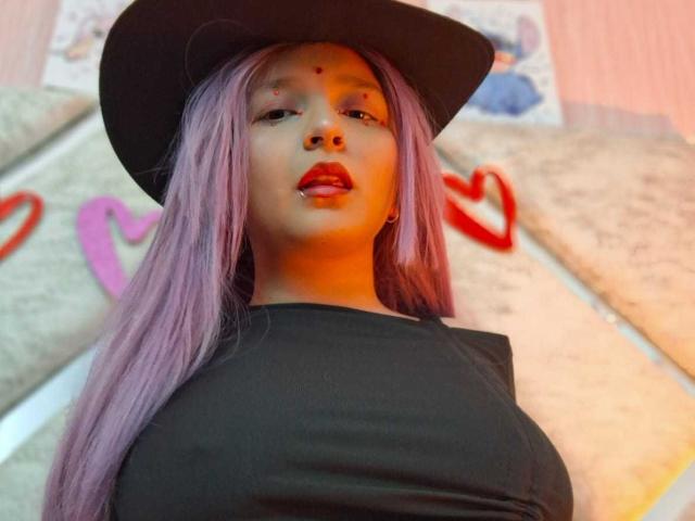CandySwan with purple hair, black hat, and black top, showing off her big tits, red lipstick, and a nose piercing, looking into the camera.
