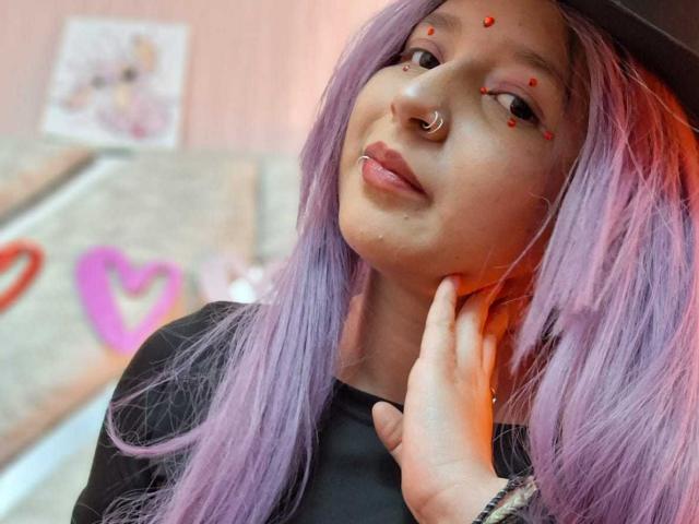 CandySwan with purple hair, nose ring, and red eye dots, touching her cheek with a hand, wearing a black top, looking at the camera, slightly pouty.