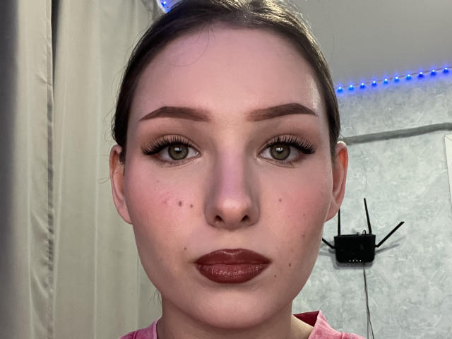 Het gezicht van JazminMensinger, met bruine ogen, donkerbruine wenkbrauwen en rode lippenstift. Ze heeft een paar moedervlekken en sproeten, en draagt lange, zwarte wimpers. Haar haar is naar achteren getrokken..