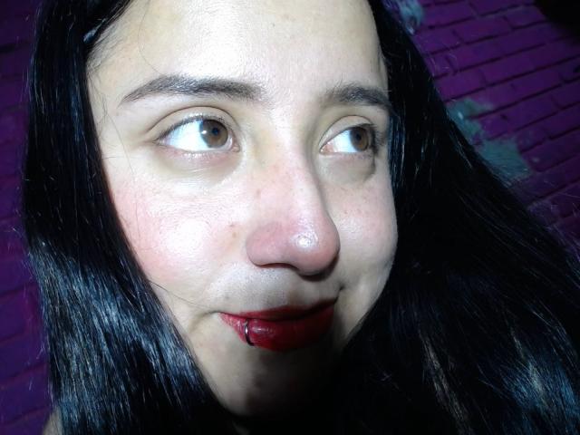 Das Gesicht von Gabriela Mora, mit braunen Augen, schwarzem Haar, rotem Lippenstift, einem leichten Lächeln und einer kleinen Nase, zeigt einige Unreinheiten und einen glänzenden, leicht feuchten Teint..