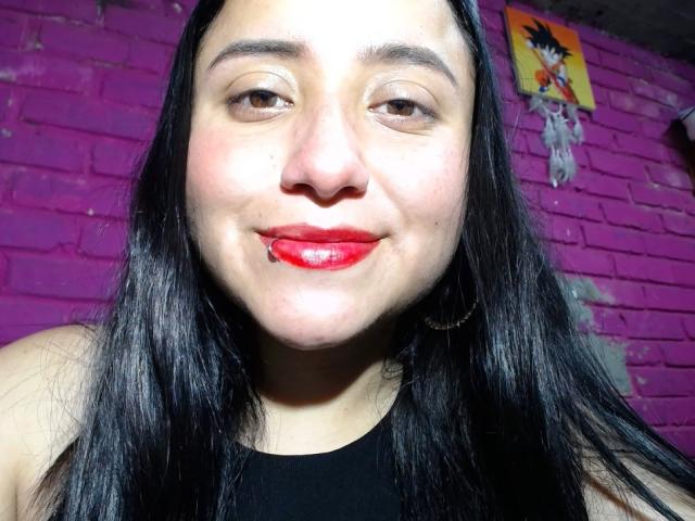 Gabriela Mora, com batom vermelho, cabelo preto e brincos de argola, sorrindo levemente com um pequeno corte no lábio inferior. Parede de tijolos roxos ao fundo..