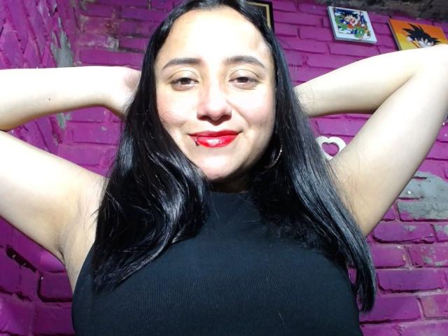 GabrielaMora com os braços levantados, blusa preta sem mangas, batom vermelho, sorrindo, cabelo preto longo, pele clara, parede de tijolos roxos, pequenas fotos emolduradas ao fundo, axila levemente visível..