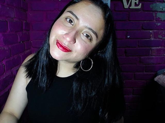 Gabriela Mora, con i capelli neri, il rossetto rosso e gli orecchini a cerchio, sorride alla fotocamera, indossando una canotta nera, ed è in piedi di fronte a un muro di mattoni viola..