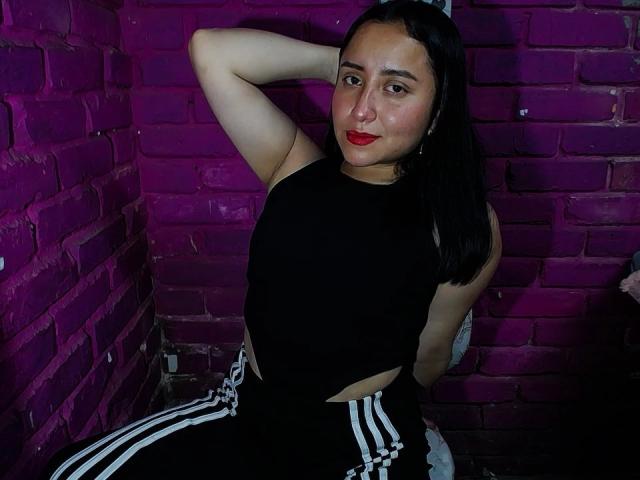 Gabriela Mora, avec du rouge à lèvres, un haut noir sans manches et un pantalon de jogging noir avec des rayures blanches, bras levé, assise contre un mur de briques roses, regardant la caméra, plan moyen..