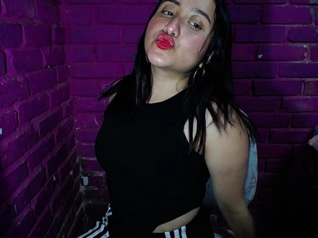Fotografia di GabrielaMora, una donna formosa con la pelle chiara e i capelli neri, che indossa un top corto nero e pantaloni a righe, con le labbra rosse e un'espressione accigliata, sullo sfondo di un muro di mattoni illuminato di viola. Incredibilmente sexy..
