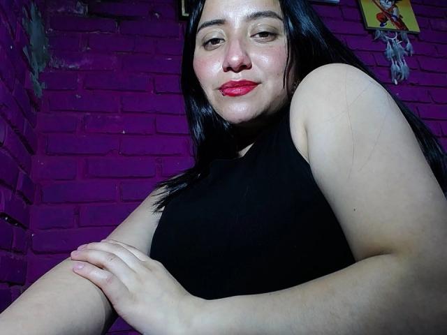 Fotografia di Gabriela Mora con lunghi capelli neri, pelle chiara e rossetto rosso, che indossa una canotta nera senza maniche, in posizione leggermente inclinata in avanti con le braccia incrociate, davanti a un muro di mattoni viola. Guarda direttamente l'obiettivo con un sorriso appena accennato..