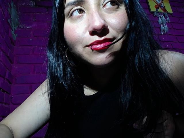 Primo piano di GabrielaMora, una donna asiatica con lunghi capelli neri, rossetto rosso e un viso leggermente arrossato, che guarda verso l'alto con un sorriso appena accennato. Sullo sfondo, un muro di mattoni viola scuro. Luce intensa sul viso..