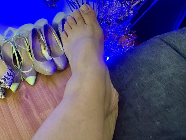 Close-up of a foot with light skin, resting next to a pile of shiny, nude-colored high heels. Foot is slightly angled, toes pointed upward. Blue and black background.