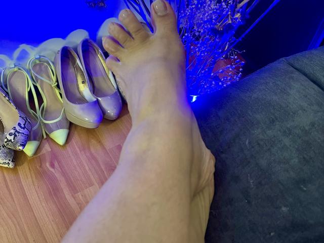 Close-up of a foot with painted toenails resting next to a pair of shiny, yellow high heels. Bright blue light glows in the background, creating a sexy, intimate atmosphere.