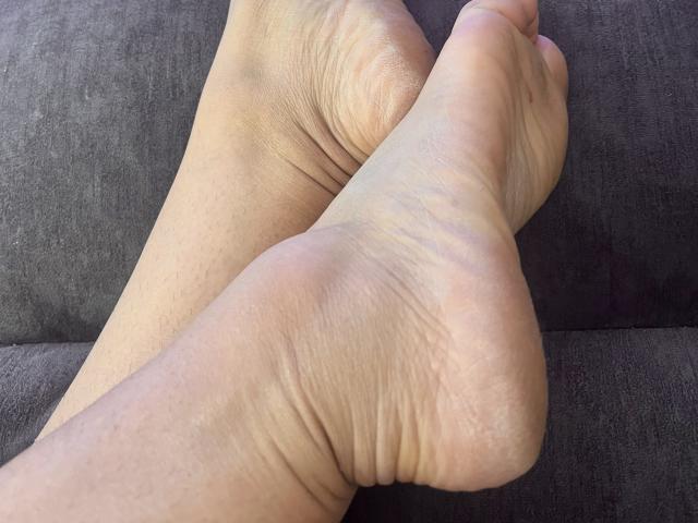 Close-up of Karimelee's right foot resting on a dark fabric surface. The foot's sole shows detailed wrinkles and slight redness, with toes pointing slightly upward. Light shadows emphasize skin texture.