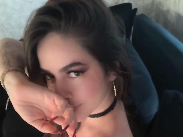 Primo piano di Christine Bray, con capelli castano scuro, che morde seducentemente il dito, indossando una collana nera e orecchini a cerchio. orecchini, i suoi occhi fissi direttamente sulla fotocamera, creando una forte tensione sessuale.