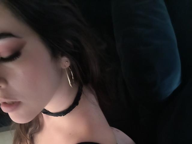 Foto em close-up de Christine Bray, com o rosto parcialmente visível, olhos fechados, usando um colar preto e um grande brinco de argola. Uma luz suave realça sua bochecha. Cabelo escuro, tensão sexual, fundo sombrio.