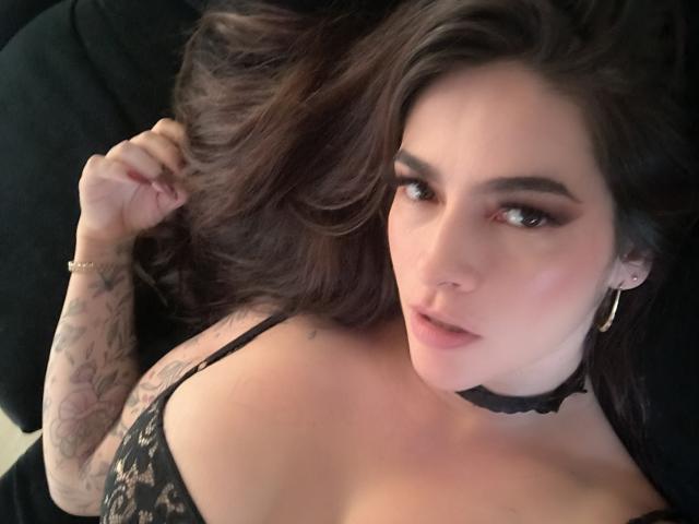 Close-up de Christine Bray, com cabelo castanho escuro, tatuagens no braço direito, vestindo uma blusa preta de renda, um colar e brincos de argola. brincos, olhando diretamente para a câmera com uma expressão sedutora..