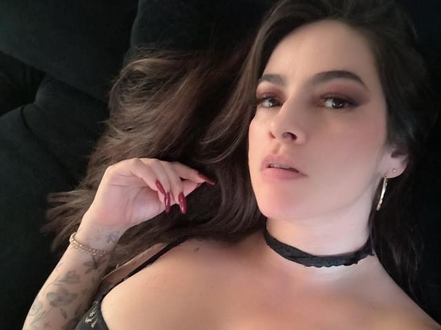 Primer plano de Christine Bray, con cabello oscuro, acostada, usando un collar negro y con un brazo tatuado. Ella está mirando seductoramente a la cámara, con uñas rojas y una expresión seria..