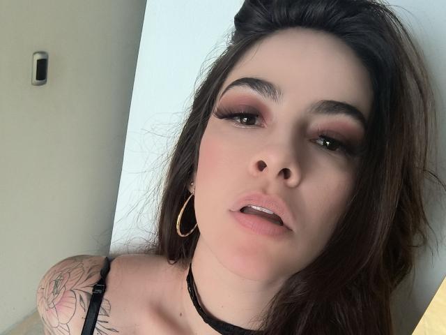 Close-up van Christine Bray, die recht in de camera kijkt met licht open lippen. Ze heeft donker haar, veel oogmake-up en een tatoeage op haar linker schouder. Draag een zwarte choker en oorringen. Lichte achtergrond..