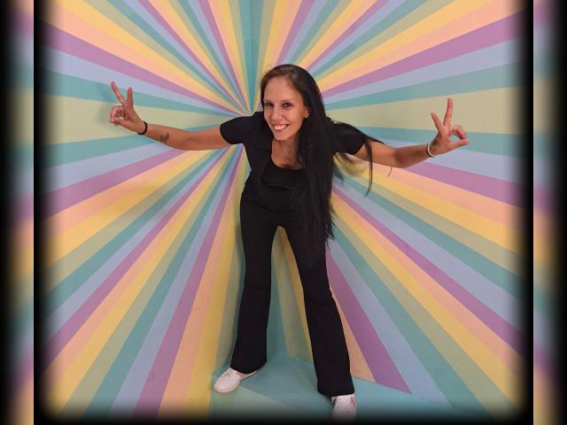 Aphrodite-88-hot, smiling with arms outstretched, flashing both middle fingers. She's wearing black clothes and white sneakers. Background: colorful, radial stripes. Bright, vibrant, and provocative. Fucking confident pose.