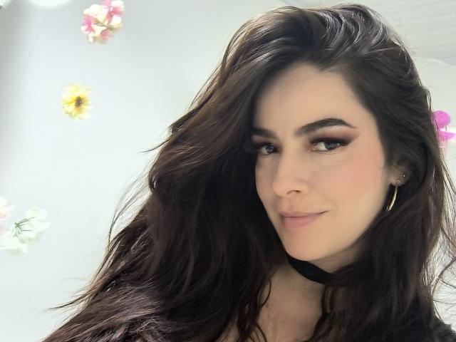 Close-up photo of ChristineBray, a young woman with long, dark brown hair, wearing a black choker, small hoop earrings, and subtle makeup. Her expression is confident, with a slight smirk. Bright background.
