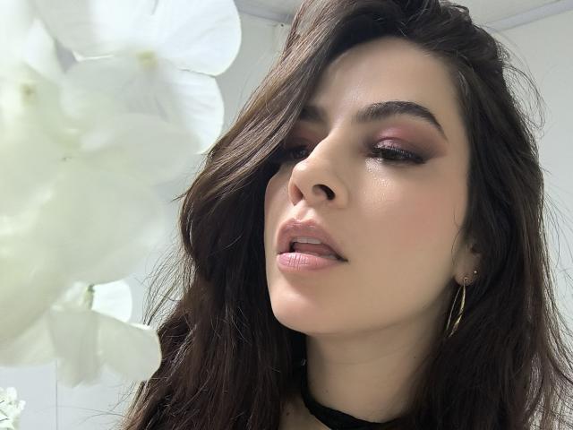 Close-up de Christine Bray, com cabelos longos e escuros, sombra rosa nos olhos e lábios brilhantes, olhando ligeiramente para o lado, com luz solar. em segundo plano, textura sutil da pele, bochechas levemente rosadas, brincos de argola..