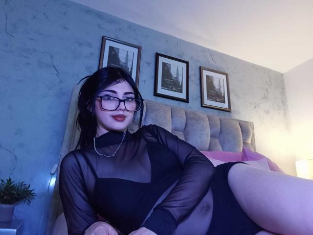 IsabellaaRossi lounges on a bed, wearing a sheer black top, exposing her tits. She has black hair, glasses, and red lipstick. Blue and white lighting highlights her sexy pose.