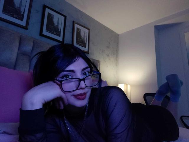 IsabellaaRossi, lying on a bed, wearing black glasses and a sheer top, head resting on hand, smiling seductively with red lips. Dark blue lighting, framed pictures on the wall, lamp in background.