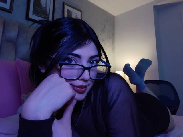 IsabellaaRossi with black hair, glasses, and red lipstick, lying on a bed, hand on cheek, legs up, wearing sheer black top, socks, dim blue lighting, seductive expression, bedroom setting.