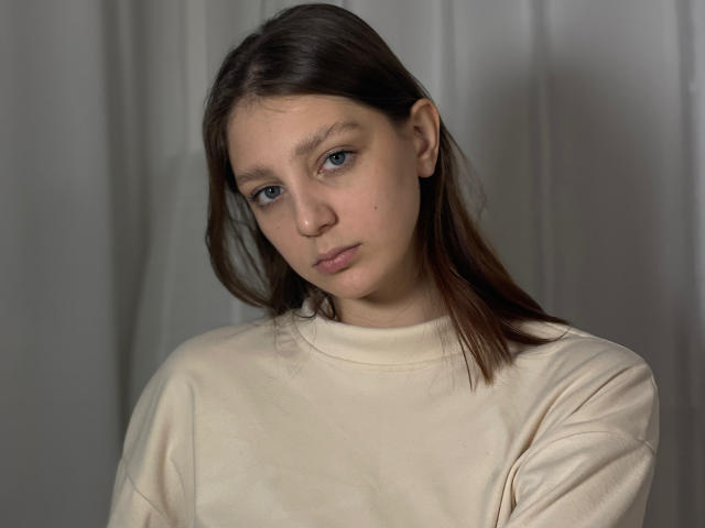 Photograph of StaceyAverbach with a serious expression, brown hair, blue eyes, wearing a plain white sweatshirt. Soft lighting, shadows on the right, gray background. Frowning, slight head tilt.