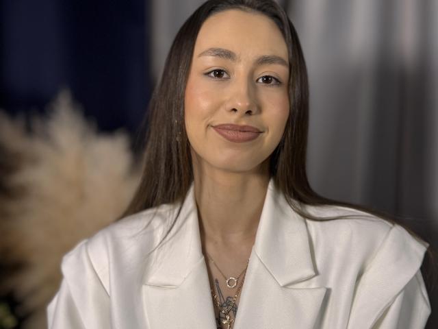 Foto van Keisha Renaker, die vooruit kijkt, gekleed in een wit blazer, met donkerbruin haar, een lichte glimlach, bruine ogen, subtiel. Blush, donkere lipstick, ketting met hanger, zachte verlichting, donkere achtergrond. Heel erg aantrekkelijk..