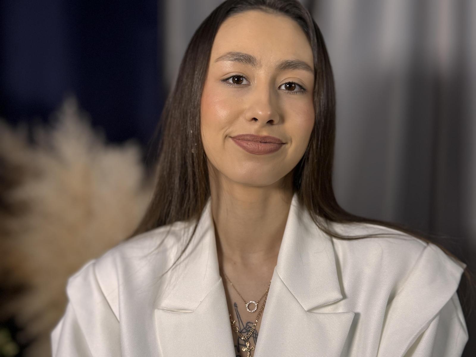 Fotografie von Keisha Renaker, die frontal aufgenommen wurde, mit einem weißen Blazer, dunklem, braunen Haar, einem leichten Lächeln und braunen Augen. Rötung, dunkler Lippenstift, Halskette mit Anhänger, weiches Licht, dunkler Hintergrund. Verdammt heiß.