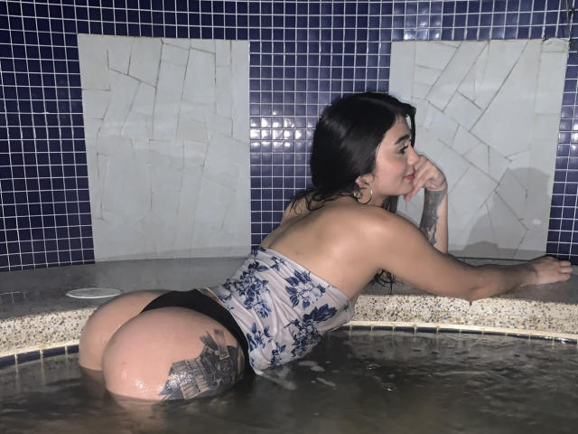 Seleneele yace boca abajo en una piscina, con las nalgas levantadas, vistiendo una blusa blanca con estampado floral y sin tirantes, y una tanga negra. Tatuaje en la mejilla derecha de su glúteo. Fondo de pared de azulejos azul oscuro..