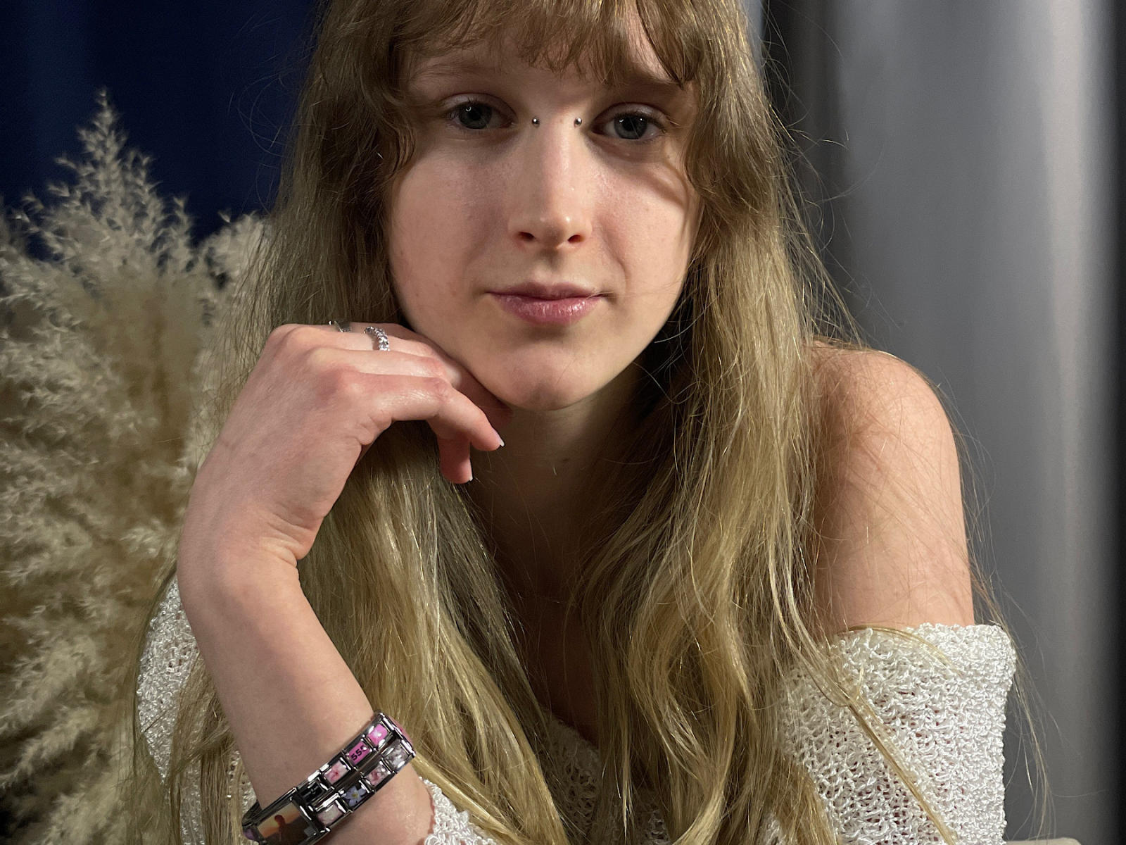Fotografia em close-up de ToiHeaslet, uma mulher branca com longos cabelos castanhos ondulados, vestindo uma blusa branca de renda com um ombro descoberto. Descansando o queixo na mão, olhando diretamente para a câmera. Pulseira visível no pulso.