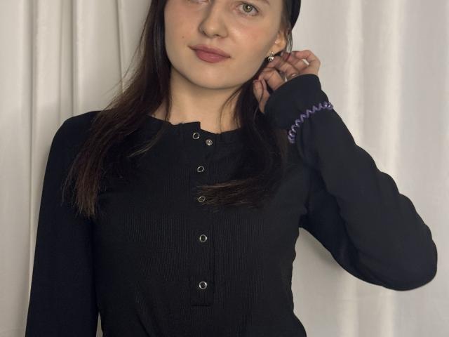 DollieSkweres, con la pelle chiara, lunghi capelli castano scuro, indossa una camicia nera a coste con bottoni, una mano tocca l'orecchio, in modo discreto. sorriso, luce naturale soffusa, sfondo con tenda bianca, leggera tensione sessuale.