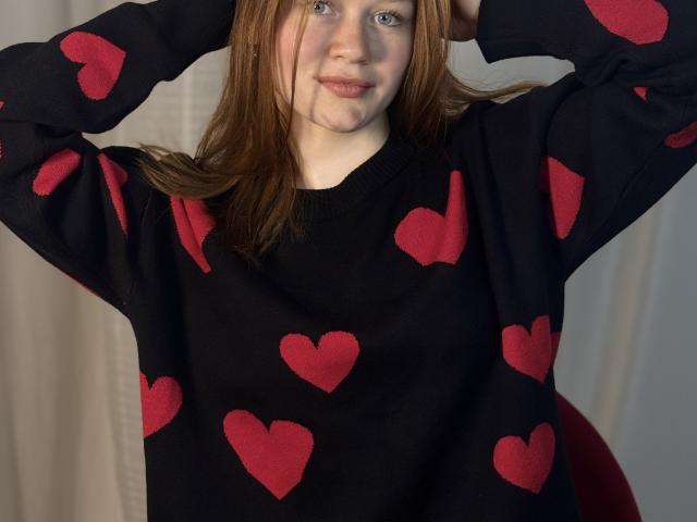 AminaEirls está de pie con los brazos levantados, vistiendo un suéter negro con diseños de corazones rojos. Ella tiene piel clara, cabello castaño y una sonrisa sutil. La imagen tiene una tensión sexual, insinuando atractivo..
