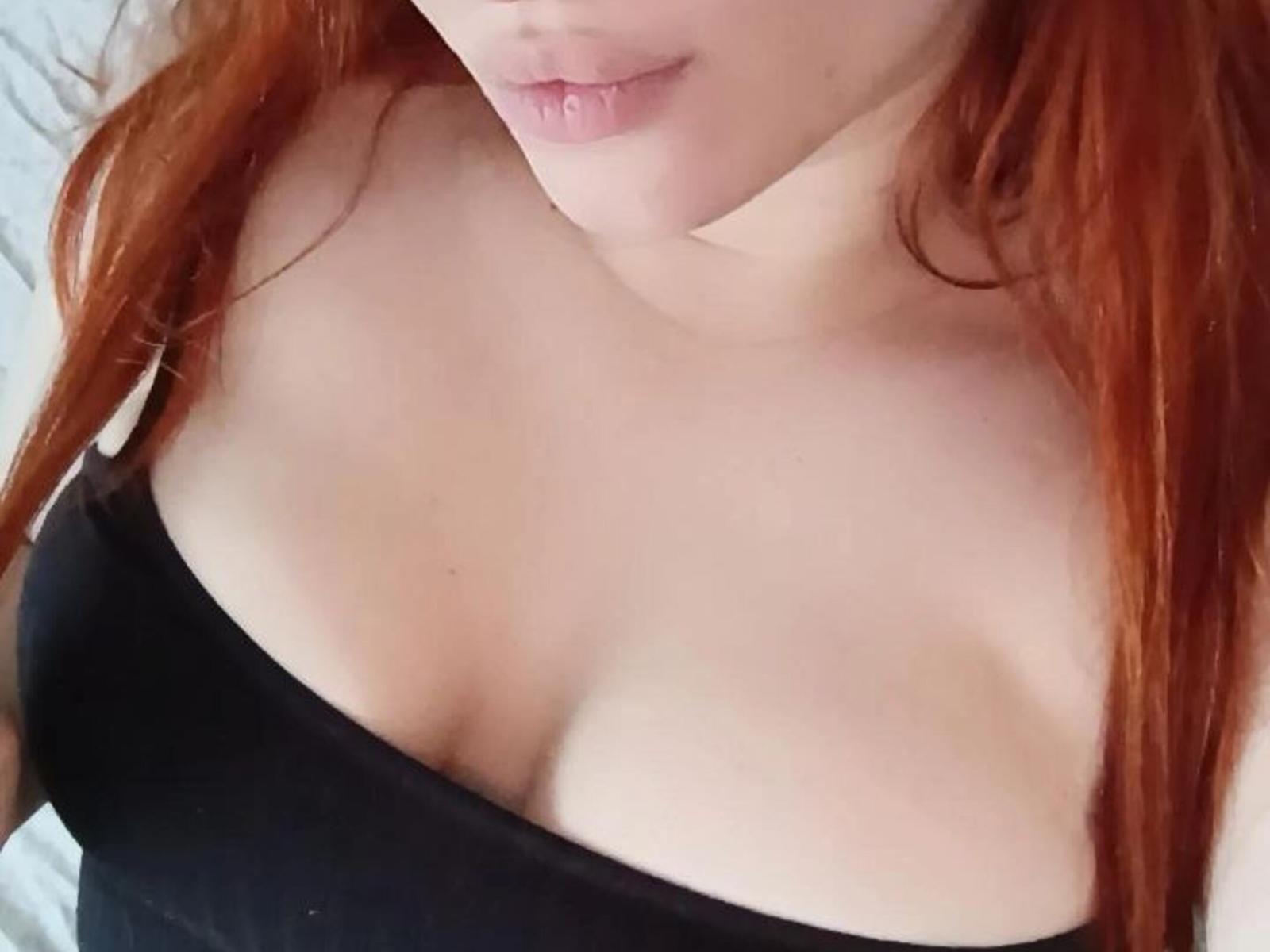Nahaufnahme von Lieselotte Mudgets blasser Haut und rotem Haar, wobei ihr Dekolleté in einem schwarzen Top zu sehen ist. Ihre Lippen sind leicht geöffnet, was eine sinnliche Spannung erzeugt. Sichtbare Schlüsselbeine und weiches Licht..