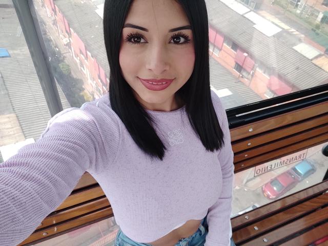 EmilyVv, com cabelos longos e pretos, vestindo uma blusa cropped branca de tricô, sorrindo para a câmera, sentada em um banco de madeira, com luz do sol. Luz entrando por uma janela, rua da cidade lá fora. Muito fofo.