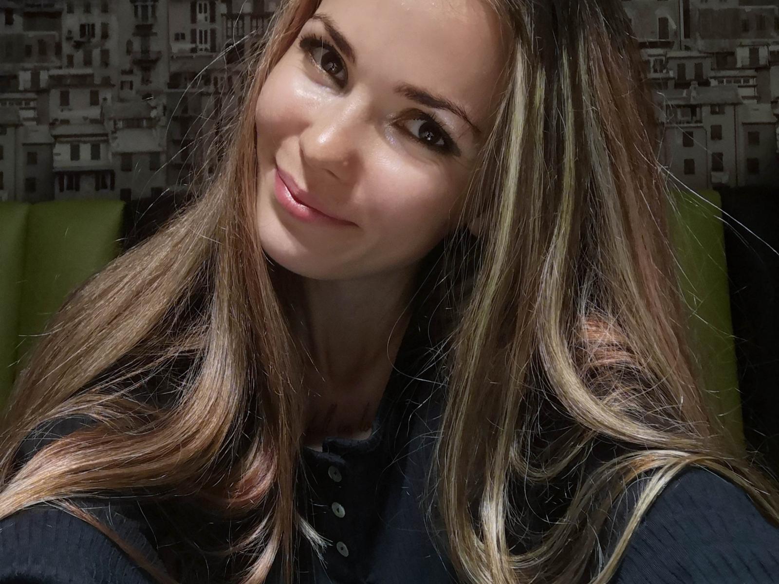Close-up de LautenSchlage com cabelo castanho longo e reto, sorrindo levemente, vestindo uma blusa preta. A luz suave realça seu rosto, criando sombras sutis. O fundo é um padrão de paisagem urbana desfocada. Muito atraente.