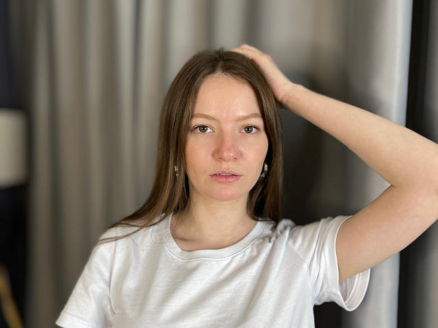 Photograph of NatishaWorthy with long brown hair, wearing a white t-shirt, hand raised behind her head, direct gaze, freckles on face, slightly parted lips, soft lighting, neutral background.