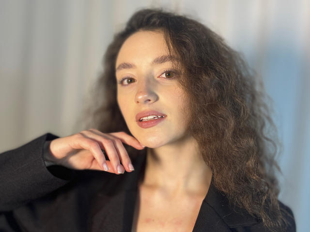 Close-up van MieshaKamimura met lang, krullend bruin haar, lichte huid en een sexy, zelfverzekerde uitdrukking. Ze draagt een zwarte top, met één hand in de buurt van haar gezicht, en het zonlicht accentueert haar gezichtsvorm..