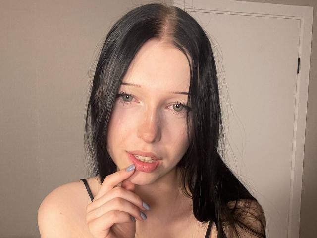 Fotografia de AmaiRin, uma jovem com pele clara e cabelos longos e pretos, mordendo o dedo de forma sedutora. Ela tem olhos azul claros e veste uma blusa preta com alças finas. Muito quente.