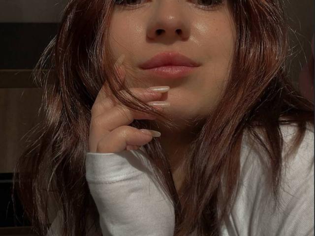 Close-up foto van BrennaMruczek, met de focus op haar gezicht en hand. Ze heeft lang bruin haar, een licht glanzende huid, volle lippen en een wit shirt met lange mouwen. Ze leunt haar kin op haar hand en kijkt recht in de camera met een subtiele, aantrekkelijke uitdrukking..