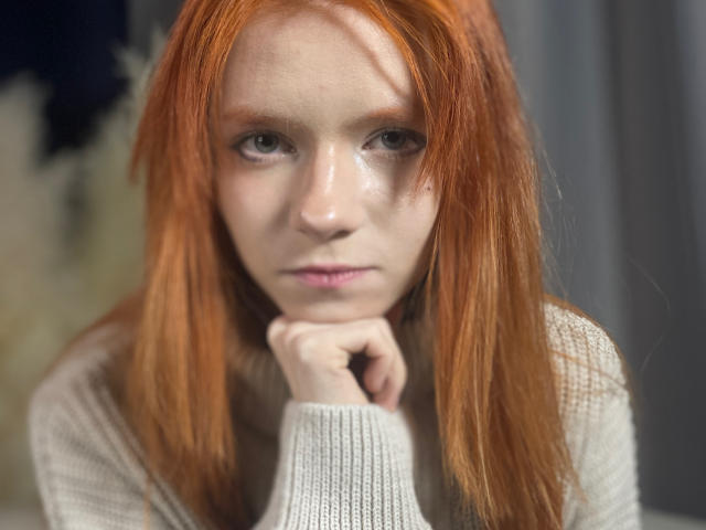 Close-up photo of JamieLathrum, a fair-skinned woman with long red hair, gazing intently with blue eyes, hand under chin, wearing a white knit top, creating intense, sexual tension.