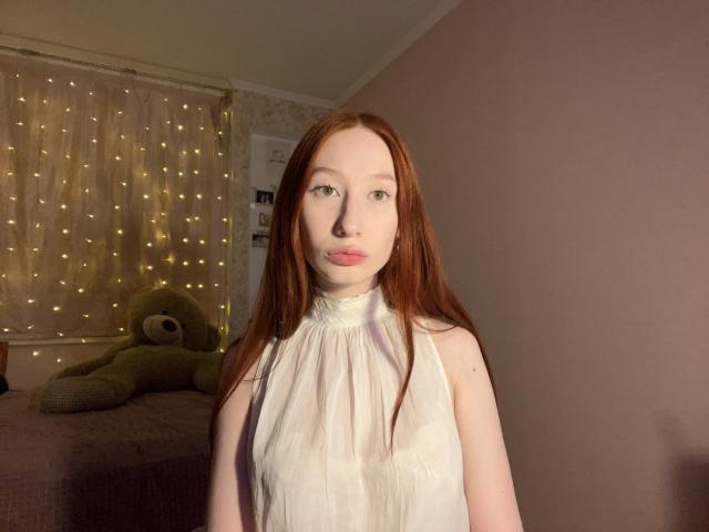 TrinhDancer, hellhäutig mit langen roten Haaren, trägt ein weißes Top ohne Ärmel und steht in einem schwach beleuchteten Raum mit Feen. Lichter und ein großer, grüner Teddybär auf einem Bett. Verdammt heiß..