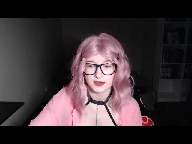 Arte digitale di LunaCherry con capelli rosa, occhiali neri e una maglietta rosa, che guarda direttamente verso la telecamera. un sorriso enigmatico.
una luce intensa illumina il suo viso, sfondo scuro..