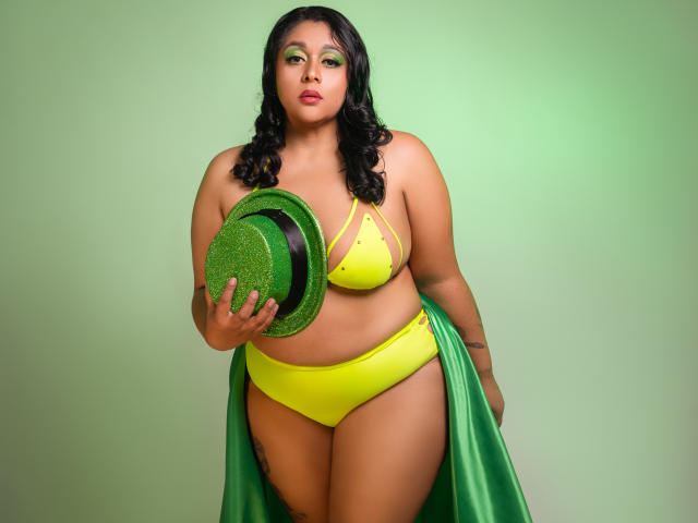 ViickyLee steht in der Mitte, kurvig und wohlgeformt, trägt einen gelben Bikini und einen grünen Umhang und hält einen grünen, glitzernden Hut. Der grüne Lidschatten passt zu ihrer selbstbewussten, verführerischen Pose. Helle, grüne Hintergrundfarbe..