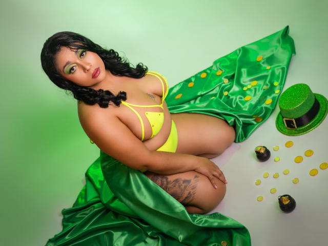 ViickyLee ajoelha-se sobre um tecido verde com padrão de trevo, vestindo um biquíni amarelo que realça seus grandes seios. Um chapéu verde e moedas de ouro estão ao lado dela. Seu cabelo preto é encaracolado..