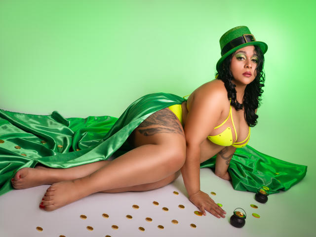 ViickyLee, a curvy woman with dark skin and black hair, lies on a green satin sheet, wearing a green hat and yellow bikini. Scattered gold coins and a black cigar nearby.