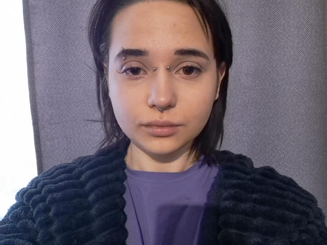 Close-up of KenyattaConsigli, a young woman with light skin, dark eyebrows, and a nose piercing. She has straight, dark hair, wears a black, textured sweater over a blue shirt. Neutral expression.