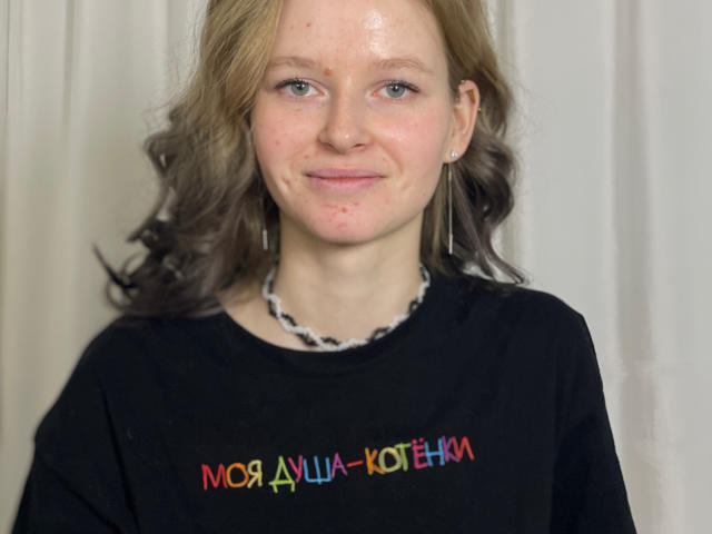 Photograph of CarlottaRenzelman with fair skin, light brown wavy hair, wearing a black shirt with colorful Cyrillic text, necklace, and earrings. She's smiling softly, displaying subtle sexual tension.