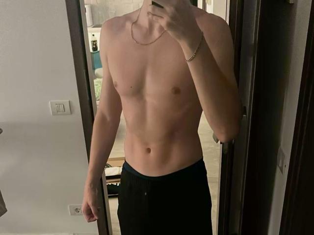 Shirtless photo of CharliePassion, slim with defined abs, wearing black pants, holding a phone to take a mirror selfie, wearing a gold necklace, dimly lit room, visible door frame.