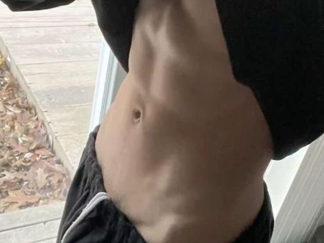 Close-up foto van het middelgedeelte van CharliePassion, waarop een getoonde, bleke buik te zien is met een duidelijke navel. CharliePassion tilt zijn zwarte shirt omhoog, waardoor zijn gespierde buik zichtbaar wordt. De achtergrond bevat een raam en een gebied met bladeren..